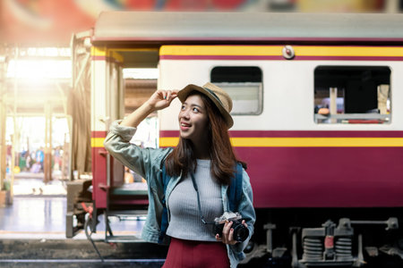 Smiling happy young Asian backpacker female standing at train station with sunshine effect. Travel...