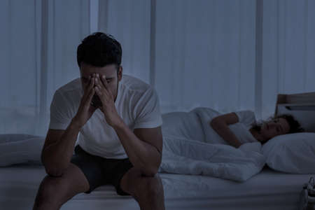 Upset depressed young man sitting on the edge of bed against his wife lying on the bed. Relationship difficulties , Problems couple.の写真素材