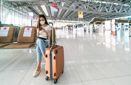 Asian female tourist wearing mask using mobile phone searching airline flight status and sit social distancing chair in airport during coronavirus or covid-19 virus outbreak a new normal conceptの写真素材