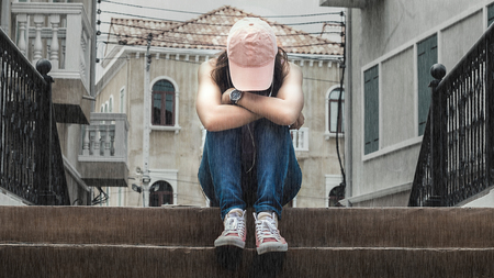 sad woman wearing a hat and hugging knees on stair in the rain