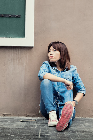 Asian woman sitting against a wall and listening to musicの写真素材