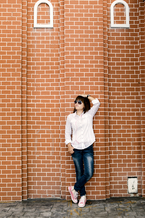 Asian woman in white shirt and jeans wearing sunglasses posing by the wall, listening to musicの写真素材