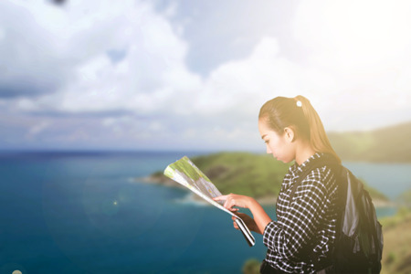 Asian woman carrying a backpack and monitoring tourist map in hand on blue sky and sea and looking...