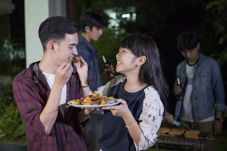 Asian young couple enjoying a romantic dinner and  group of friends having outdoor garden barbecue laughing with alcoholic beer drinks on nightの写真素材