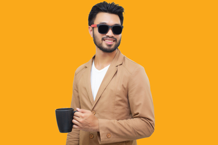 Portrait of a handsome asian young businessman holding an cup and drinking coffee. Isolated on yellow background.の写真素材