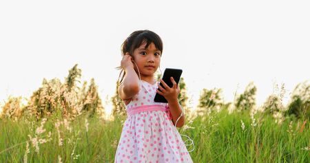 Asian child girl Listening to music and singing from a mobile phone and happy on meadow in summer in natureの写真素材