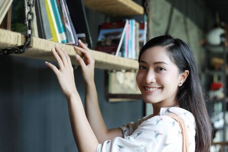 A cute Asian woman choose a book on the bookshelf for reading and she smiled happy on her holiday at home.の写真素材