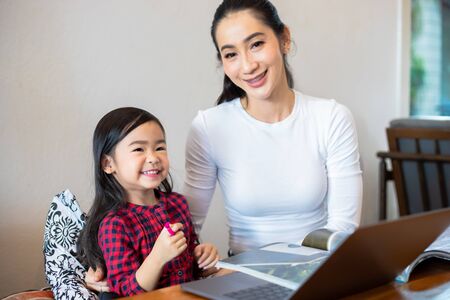 Asian mothers are teaching their daughters to read a book and use notebooks and technology for online learning during school holidays at home. Educational concepts and activities of the familyの写真素材