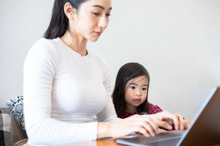 Asian mothers are teaching their daughters to read a book and use notebooks and technology for online learning during school holidays at home. Educational concepts and activities of the familyの写真素材