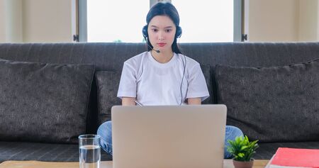 Asian happy woman student wear headphones study online watching webinar podcast on laptop listening learning education course conference calling and write notes look at laptop sit on sofa at homeの写真素材