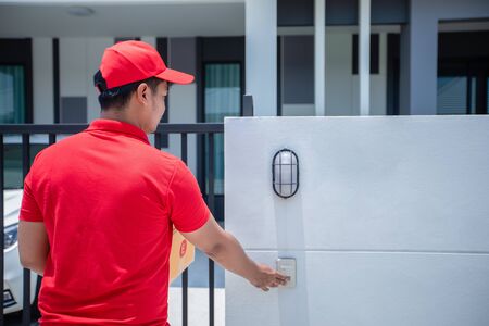 Asian delivery servicemen wearing a red uniform with a red cap handling cardboard boxes to give to the customer in front of the house and Click the doorbell. Online shopping and Express deliveryの写真素材