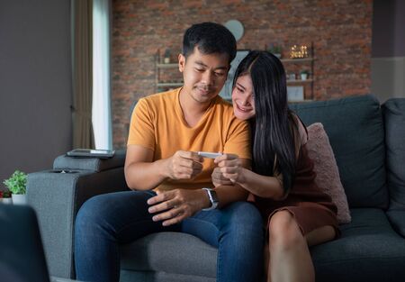 Happy Asian couple smiling looking cheerful after a home pregnancy test in sofa at homeの写真素材