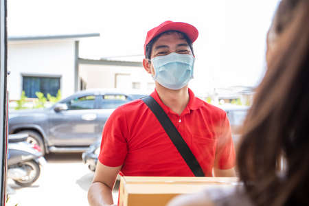 Asian delivery servicemen wearing a red uniform with a red cap and face mask handling cardboard boxes to give to the female customer in front of the house. Online shopping and Express delivery