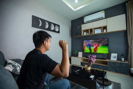 Asian man is watching football on tv and sit on the sofa at home on a holiday and smiling and laughing happy.の写真素材