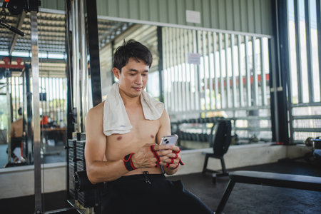young male playing phone and listening to music after exercise with various exercise equipment in...