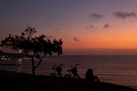 The backlight of the tree, bicycles and people creates a context of protection, freedom, intimacy and individuality that stands out in this photograph related to love and friendship. Backlight of a couple watching the sunset from the Lima boardwalk.の写真素材