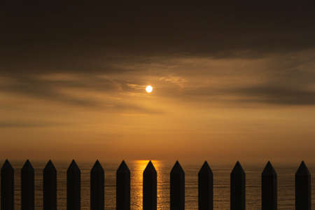 Sun behind the bars of a fence. Sunset with the sun on the horizon, behind stake gates.