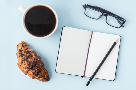 Office blue table with blank notepad, pencil, glasses, fresh croissant and coffee cup. Copy space for text. Flat lay. Top viewの写真素材