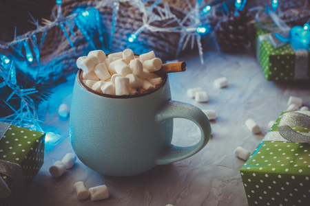 Christmas and New Year cozy holiday composition with gift boxes, lights, cinnamon, scarf, pine cone, mugs with cocoa or chocolate, marshmallow on the gray concrete background.の写真素材
