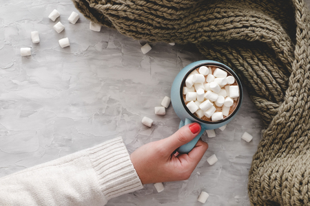 Christmas and New Year cozy holiday composition with scarf, woman hand, mugs with cocoa or chocolate, marshmallow on the gray concrete background. Flat lay, top view.の写真素材