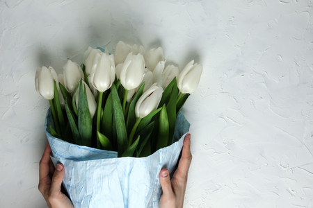 A bouquet of white tulips in blue wrapping paper with woman hands on a white concrete background. Top view. Flat lay. Postcard for Easter, Mothers Day and Spring Holidays.の写真素材