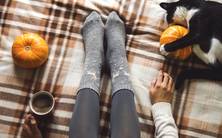 Womens hands and feet in sweater and woolen cozy gray socks holding cup of hot coffee, sitting on plaid with kitten, pumpkin, candles and leaves. Concept winter comfort, morning drinking.の写真素材