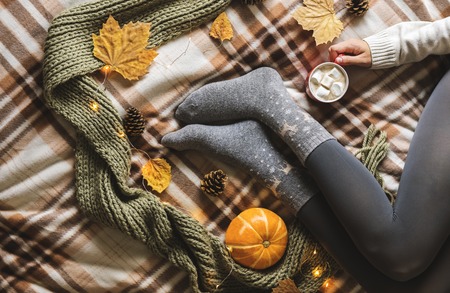 Womens hands and feet in sweater and woolen cozy gray socks holding cup of hot coffee with marshmallow, sitting on plaid with pumpkin, knitted scarf, leaves. Concept winter comfort, morning drinking.の写真素材