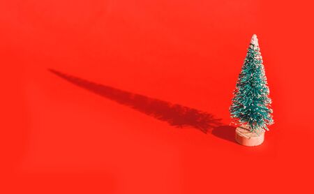 Miniature toy artificial Pine tree on wooden base on bright red Christmas background with long shadow. Concept of New Year, Christmas holiday greeting card.の写真素材