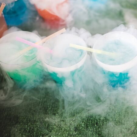 Steaming multi-colored cocktails with dry ice standing on green background.