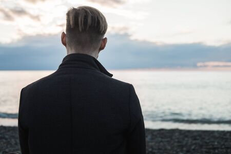 Young male hipster in a coat sits by the sea or ocean and enjoys sunset. Autumn by the sea on a rocky beachの写真素材