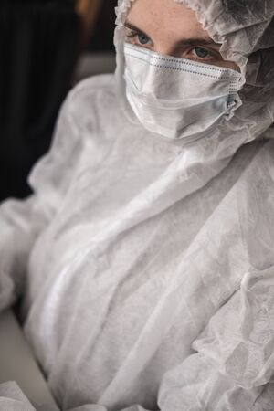Girl in medical mask and white transparent protective suit sits in quarantine at home and works at...