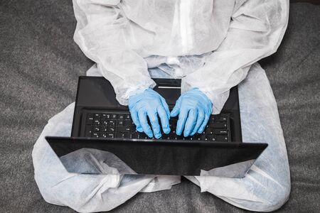 Person in protective white translucent suit, blue rubber gloves, medical mask sits at table at home and works or studies on laptop during quarantine. Remote work during coronavirus pandemicの写真素材