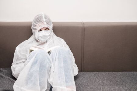 Girl in protective white translucent suit, blue rubber gloves, a medical mask sits on sofa at home and reads book. Stay home during the coronavirus pandemicの写真素材