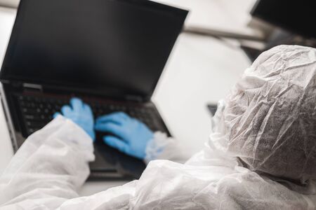 Person in protective white translucent suit, blue rubber gloves, medical mask sits at table at home and works or studies on laptop during quarantine. Remote work during coronavirus pandemic.の写真素材