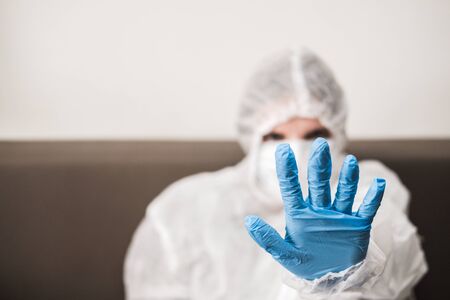 Person in protective white translucent suit, blue rubber gloves, medical mask extends his hand forward. Gesture stay home during the coronavirus pandemic.の写真素材