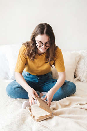 Young beautiful girl student in glasses, an orange T-shirt and blue jeans sits on bed with pillows...
