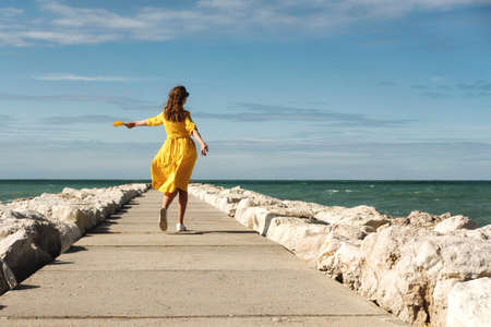 Young beautiful woman in a bright yellow dress stands on the pier on the seashore or ocean with her arms wide open. Concept of freedom, enjoyment of nature, thoughtfulness, mindfulness.の写真素材