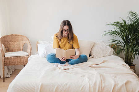 Young beautiful girl student in glasses sits on bed with pillows and reads paper book.の写真素材