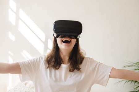 Young woman in jeans and white T-shirt with virtual reality glasses on her head moves her arms,...