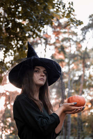 Young woman in dark dress and witch hat holds a pumpkin in her hands. Halloween party costume. Forest, park with autumn trees.の写真素材