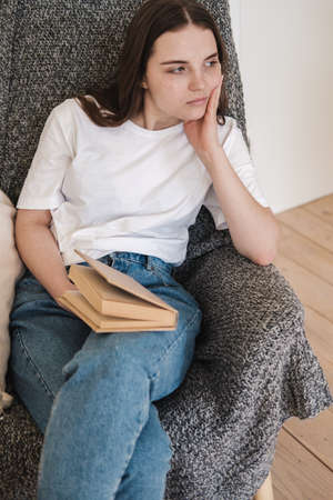 Young woman in jeans and white T-shirt sits in gray armchair and reads paper book. Concept of relaxing holiday, home comfort, hygge, slow-living.の写真素材