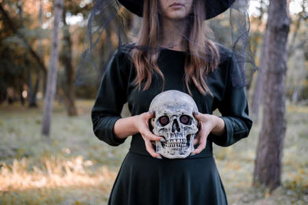 Young beautiful cute woman in dark dress and witchs hat holds a skull in her hands. Halloween party costume. Forest, park with autumn trees.の写真素材