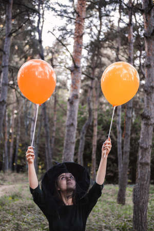 Young cute beautiful woman in dark dress and witchs hat holds orange balloons in her hands. Halloween party costume. Forest, park with autumn trees.の写真素材