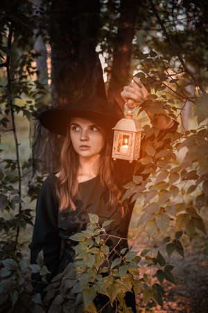 Young woman in dark dress and witchs hat holds lantern with candles in her hands and illuminates the...