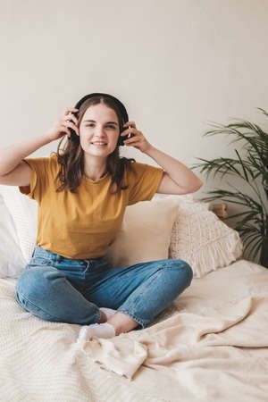 Young beautiful girl student in an orange T-shirt and blue jeans sits on bed with pillows and listens to an audiobook, music, podcast on headphones. Self-education concept at home during quarantine.の写真素材