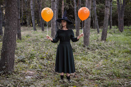 Young cute beautiful woman in dark dress and witchs hat holds orange balloons in her hands....