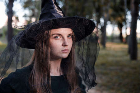 Pretty young beautiful woman in dark dress and witch hat standing in the middle of the autumn woods or park. Halloween party costume. Close-up portrait. Shadow on her face.の写真素材