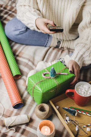 Female hand with smartphone takes pictures of New Years or Christmas flatlay for social networks. Creating content. Wooden tray with cocoa mug marshmallows, toy tree, candle, stars, wrapped gift.の写真素材