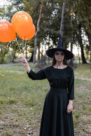 Young cute beautiful woman in dark dress and witchs hat holds orange balloons in her hands....