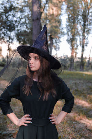 Pretty young beautiful woman in dark dress and witch hat standing in the middle of the autumn woods or park. Halloween party costume. Close-up portrait. Shadow on her face.の写真素材
